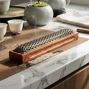 May include: A rectangular wooden incense burner with a gold-colored, lattice-patterned top. Wisps of smoke rise from the burner, suggesting the use of incense. The burner sits on a patterned mat, with teacups and a small potted plant in the background.