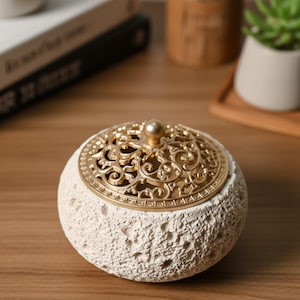 Op de afbeelding: Een ronde, crèmekleurige wierookbrander met een getextureerd oppervlak. De goudkleurige deksel heeft een ingewikkeld, opengewerkt ontwerp en een kleine knop. Een decoratief item.