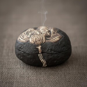 Può includere: Un porta incenso in ceramica nera con un motivo floreale dorato sul coperchio. Vapore sale dal porta incenso, suggerendo l'uso di incenso. Il porta incenso rotondo è posto su una superficie strutturata.
