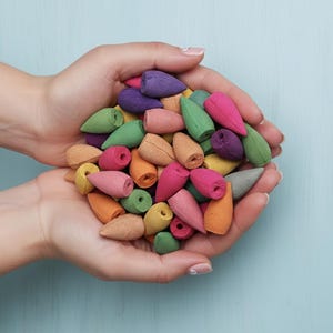 以下が含まれることがあります： ピンク、緑、紫、オレンジ、黄色など、さまざまな色のカラフルなお香コーンのコレクション。お香コーンは、明るい青色の背景に対して、すぼまった手の中に保持されています。コーンは円錐形をしています。