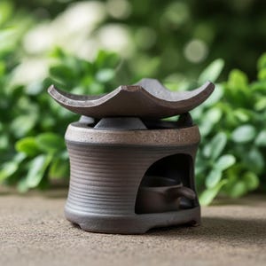 May include: A brown ceramic essential oil diffuser with a wavy top and a small opening for a tea light. The diffuser has a ribbed body and a small cup inside. The background is blurred green foliage.