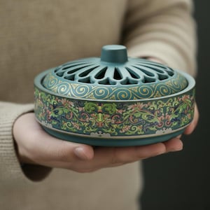 May include: A teal ceramic incense burner with a lid featuring a series of arched cutouts. The body is adorned with a detailed floral and scrollwork pattern in shades of green, pink, and gold. The burner is held in open hands.