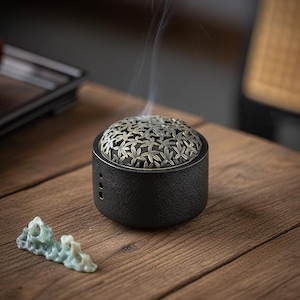 Könnte beinhalten: Ein schwarzer Räuchergefäß mit einem dekorativen Deckel mit Blättermuster. Rauch steigt aus dem Brenner auf, was auf die Verwendung hindeutet. Ein kleines, hellgrünes Dekorationsteil liegt in der Nähe auf einer Holzoberfläche. Der Brenner hat eine strukturierte Außenseite.