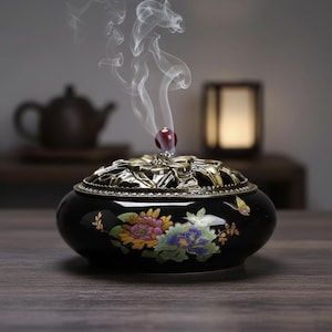 Könnte beinhalten: Ein schwarzer Keramik-Räuchergefäß mit einem dekorativen Blumendesign in Gelb, Rosa und Blau. Der Brenner hat einen goldfarbenen, durchbrochenen Deckel und eine rote Perle oben. Rauchschwaden steigen aus dem Brenner auf, was darauf hindeutet, dass er in Gebrauch ist.