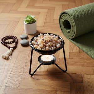 Op de afbeelding: Een zwarte metalen wierookbrander met een brandende kaars, gevuld met amberkleurige hars. Een groene yogamat, een kleine vetplant in een witte pot, een gebedsketting en twee grijze stenen zijn ook aanwezig.