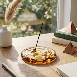 Op de afbeelding: Een ronde wierookhouder van amberkleurig glas met een golvende, waterachtige oppervlaktestructuur. Eén dun zwart wierookstokje staat rechtop in het midden van de houder en verspreidt een dun sliertje rook in de lucht.