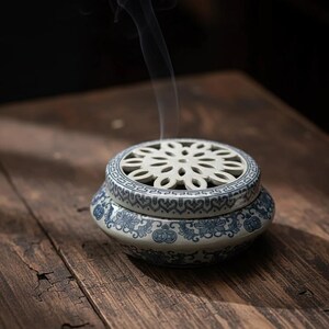 May include: A ceramic incense burner with a white floral lid and blue floral patterns on the base. Smoke rises from the burner, indicating incense use. The burner is placed on a wooden surface.