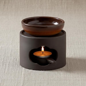 Puede incluir: Un quemador de aceite de cerámica marrón oscuro con un cuenco pequeño y redondo de color marrón en la parte superior. Una vela de té encendida está dentro del quemador, visible a través de una abertura decorativa. El quemador está sobre una superficie neutra.