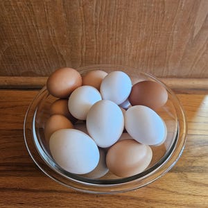 May include: A clear glass bowl filled with a mix of brown and white eggs. The eggs vary in shade from light tan to dark brown. The bowl sits on a wooden surface, with a wooden backdrop.