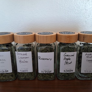 May include: Five glass jars with wooden lids filled with dried herbs. The labels read: Basil, Ground Lemon Balm, Rosemary, Ground Apple Mint, and Apple Mint Leaves. Each jar has a date on the label.