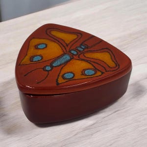 May include: A triangular ceramic trinket box with a deep reddish-brown base and a lid featuring a stylized butterfly design. The butterfly is rendered in shades of golden yellow and bright blue, outlined in dark brown, creating a contrast.