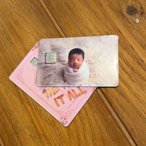 May include: Two rectangular plastic cards with rounded corners. The top card features a photograph of a baby wrapped in a white blanket on a fluffy surface and a gold chip. The bottom card is pink with gold text reading HE HAS IT ALL.