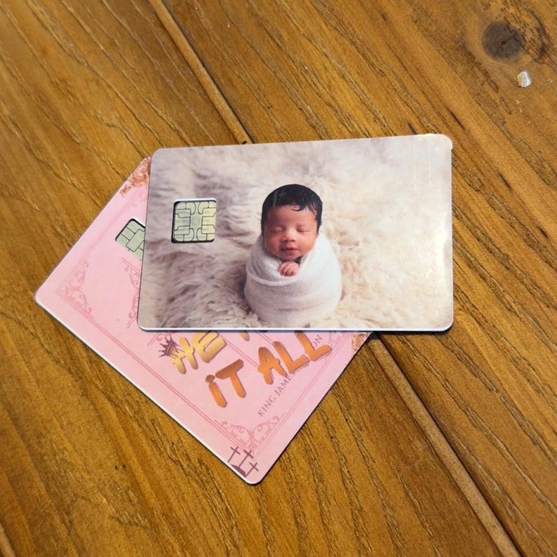 May include: Two rectangular plastic cards with rounded corners. The top card features a photograph of a baby wrapped in a white blanket on a fluffy surface and a gold chip. The bottom card is pink with gold text reading HE HAS IT ALL.
