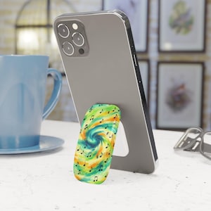 May include: A smartphone propped up by a colorful phone grip. The grip features a swirling design in shades of green, yellow, and blue, adorned with musical notes. A blue mug and artwork are in the background.