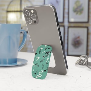 Könnte beinhalten: Ein Smartphone mit einem türkisfarbenen Pop-up-Telefonhalter mit einem Notenmuster in Schwarz und Weiß. Das Telefon ist grau und steht auf einer weißen Oberfläche. Eine blaue Tasse ist im Hintergrund.