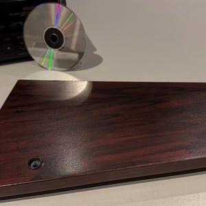 Op de afbeelding: Een donkerhouten CD-spelerbasis met twee zwarte schroefgaten. Een zilveren cd met regenboogreflecties staat op het oppervlak. Op de achtergrond staat een zwart stereocomponent.