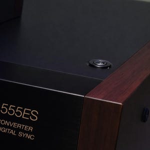 Op de afbeelding: Close-up van een zwart audio-onderdeel met een donkere houten afwerking. De tekst "555ES" is zichtbaar in gouden letters, samen met "CONVERTER DIGITAL SYNC" en "C. INDEX". Een kleine, ronde knop bevindt zich op het bovenoppervlak.