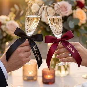 May include: Two champagne flutes clinked together, filled with a golden liquid. Each glass has a decorative bow tied around the stem. One bow is black with the word "Bräutigam" and the other is burgundy with the word "Braut".