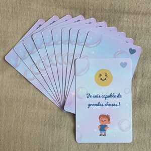 Peut inclure: Un jeu de cartes illustrées avec un motif de bulles pastel. Une carte présente un visage jaune souriant, une illustration d'enfant et le texte français "Je suis capable de grandes choses!"