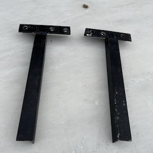 May include: Two black metal shelf brackets in a T-shape. Each bracket has a horizontal top with three holes for mounting and a vertical support. The brackets are on a light-colored surface.