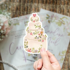 May include: A sticker of a tiered wedding cake with floral decorations and a heart on top. The cake is held in a hand, with wedding invitations and a bouquet in the background. The cake has a light beige color.