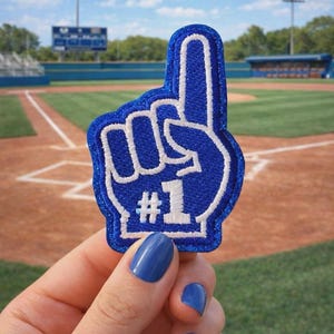 May include: A blue foam finger with a white outline and the number one. The foam finger is held up in front of a baseball field. The background includes the baseball field, bleachers, and a scoreboard.