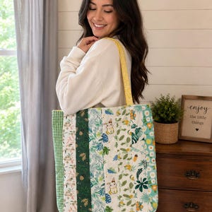 May include: A quilted tote bag featuring vertical fabric strips in various patterns, including green gingham, dark green with acorns, white with botanical leaves and small rabbits, and a light blue floral print, finished with yellow patterned shoulder straps.