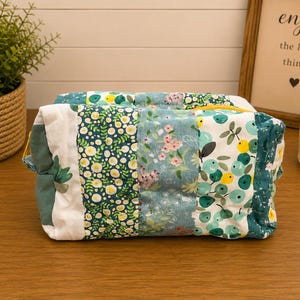 May include: A rectangular fabric pouch featuring a patchwork design of various floral and botanical patterns in shades of teal, green, white, and yellow. The quilted accessory has a yellow zip closure and a soft, padded construction with vertical panels.