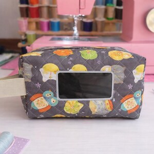 May include: A gray quilted fabric box with a rectangular window and a cream-colored handle. The box is decorated with colorful cartoon-like creatures. Sewing supplies and a pink sewing machine are in the background.