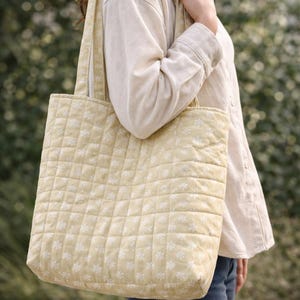 May include: A quilted tote bag in a light yellow color with a white floral pattern. The bag has a square shape and a quilted texture. The straps are the same color and pattern as the bag.