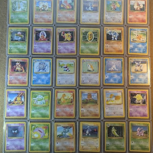 May include: A collection of vintage Pokémon trading cards, each encased in a protective sleeve. The cards feature various colorful illustrations of Pokémon characters, with text and symbols. The cards are arranged in a grid pattern.