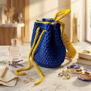 Könnte beinhalten: Ein handgefertigter blauer Häkelrucksack mit gelben Verzierungen und Trägern. Der Kordelzugbeutel wird aus recyceltem Stoffgarn hergestellt. Das Bild zeigt auch Goldschmuck, Parfüm und Make-up.