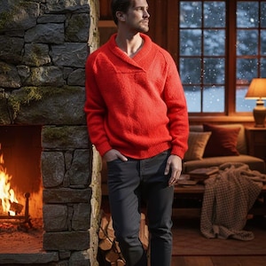 May include: A man wearing a bright red sweater with a shawl collar and dark gray jeans. He is standing near a stone fireplace with a fire burning inside. The background includes a cozy living room with a window overlooking a snowy scene.