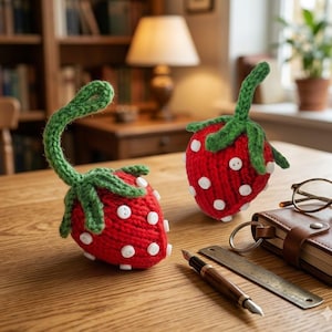 Könnte beinhalten: Zwei handgefertigte, gestrickte Erdbeeren mit grünen Stielen und Blättern sowie weißen Knopfsaaten. Die rote Frucht liegt auf einem Holztisch mit einem ledergebundenen Notizbuch, einem Stift und einer Brille. Die Erdbeeren sind etwa 7.6 cm hoch.