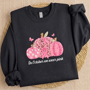 May include: A black long-sleeve sweatshirt featuring a graphic of three pink pumpkins with various patterns including leopard print, polka dots, and breast cancer awareness ribbons. A pink ribbon is centered below the pumpkins with the text In October we wear pink.