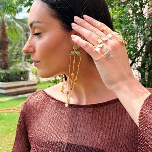 May include: A woman wearing gold earrings and rings with geometric designs. The earrings feature cascading chains with various shapes and colors. The rings have a similar design, with square and diamond-shaped accents. The woman is wearing a maroon top.