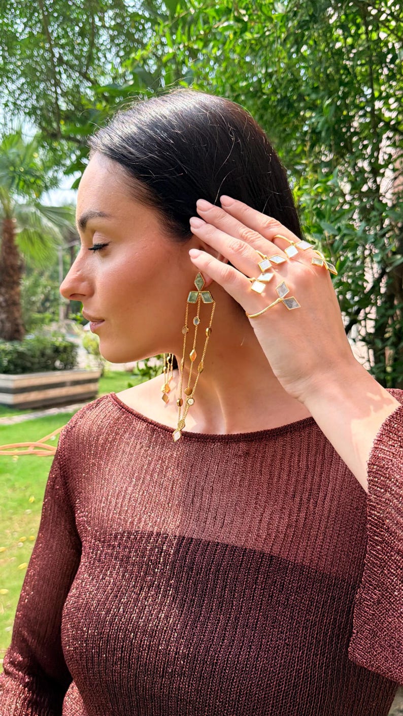 May include: A woman wearing gold earrings and rings with geometric designs. The earrings feature cascading chains with various shapes and colors. The rings have a similar design, with square and diamond-shaped accents. The woman is wearing a maroon top.