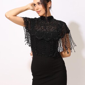 Model wearing a black embroidered beaded cape with scalloped edges and fringe. The sheer net base is hand-embellished with intricate embroidery and sparkling black beads, creating a statement bridal or evening accessory.
