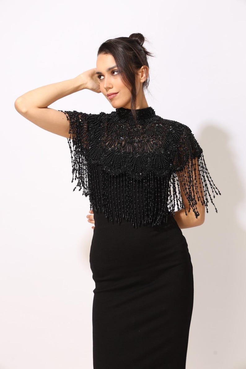 Model wearing a black embroidered beaded cape with scalloped edges and fringe. The sheer net base is hand-embellished with intricate embroidery and sparkling black beads, creating a statement bridal or evening accessory.