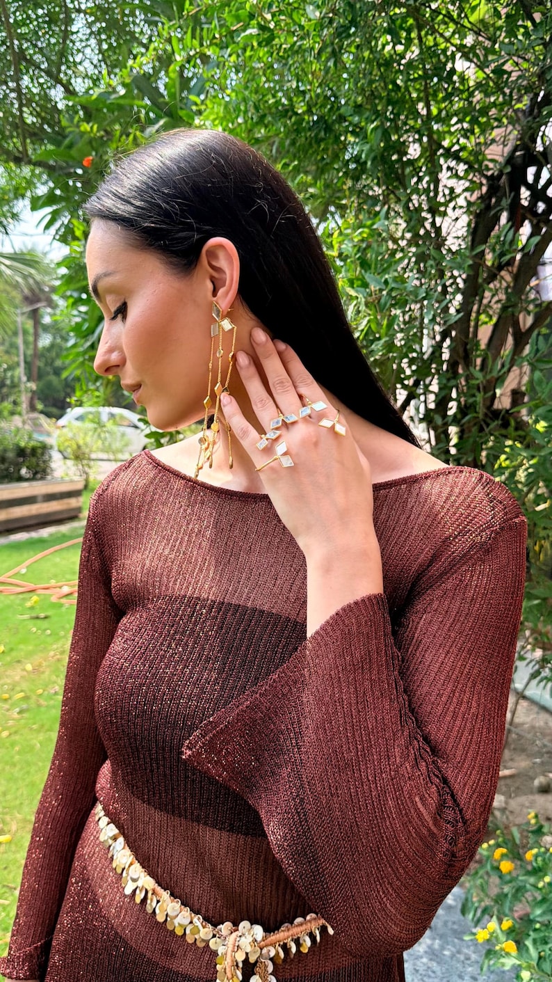 May include: A woman wearing gold earrings and rings with geometric designs. The earrings have multiple dangling chains. The rings are on multiple fingers. She is wearing a long-sleeved, sheer, brown top with a matching belt.