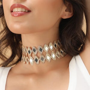 May include: A gold-toned choker necklace featuring a geometric design of mirrored and reflective diamond shapes. The necklace has a delicate, intricate pattern, with alternating mirrored and solid sections. The jewelry is worn around the neck.