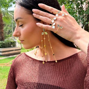May include: A woman wearing gold-toned jewelry, including a ring and earrings. The ring features geometric shapes, and the earrings have dangling chains with various beads. The woman is wearing a maroon-colored top.