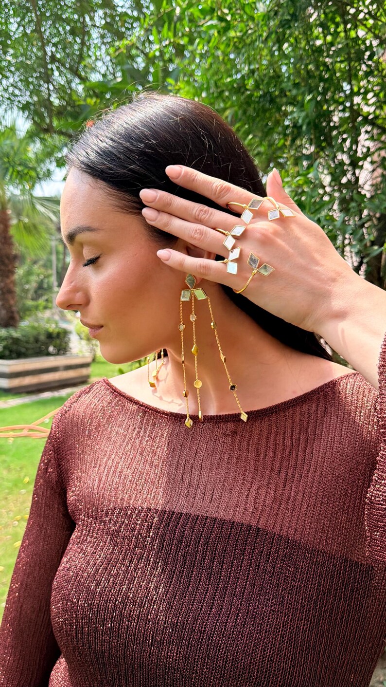 May include: A woman wearing gold-toned jewelry, including a ring and earrings. The ring features geometric shapes, and the earrings have dangling chains with various beads. The woman is wearing a maroon-colored top.