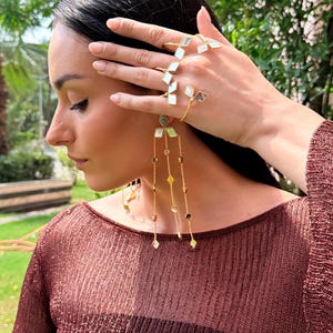 May include: Gold-toned jewelry set featuring geometric designs. The set includes rings and long dangling earrings with diamond-shaped and rectangular accents. The model is wearing a maroon top, and the background is a blurred green.
