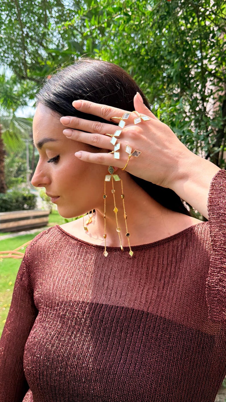 May include: Gold-toned jewelry set featuring geometric designs. The set includes rings and long dangling earrings with diamond-shaped and rectangular accents. The model is wearing a maroon top, and the background is a blurred green.