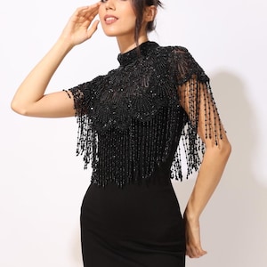 Model wearing a black embroidered beaded cape with scalloped edges and fringe. The sheer net base is hand-embellished with intricate embroidery and sparkling black beads, creating a statement bridal or evening accessory.