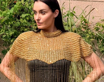 May include: A gold beaded cape with fringe detail, worn over a black dress. The cape has a high, rounded neckline and a layered design. The fringe hangs down the arms and chest. The background includes greenery and a pink wall.