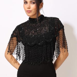 Model wearing a black embroidered beaded cape with scalloped edges and fringe. The sheer net base is hand-embellished with intricate embroidery and sparkling black beads, creating a statement bridal or evening accessory.