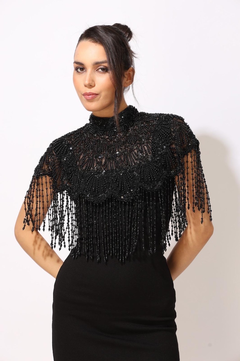 Model wearing a black embroidered beaded cape with scalloped edges and fringe. The sheer net base is hand-embellished with intricate embroidery and sparkling black beads, creating a statement bridal or evening accessory.