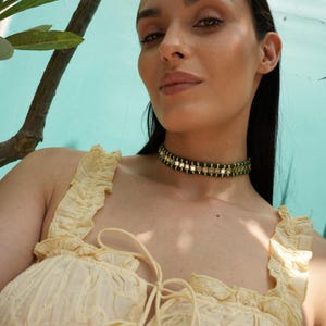 May include: A choker necklace featuring a dark green woven band accented with a row of rectangular, clear, reflective stones set in gold-toned metal. The necklace is worn with a pale yellow, sleeveless, textured top with ruffled straps.